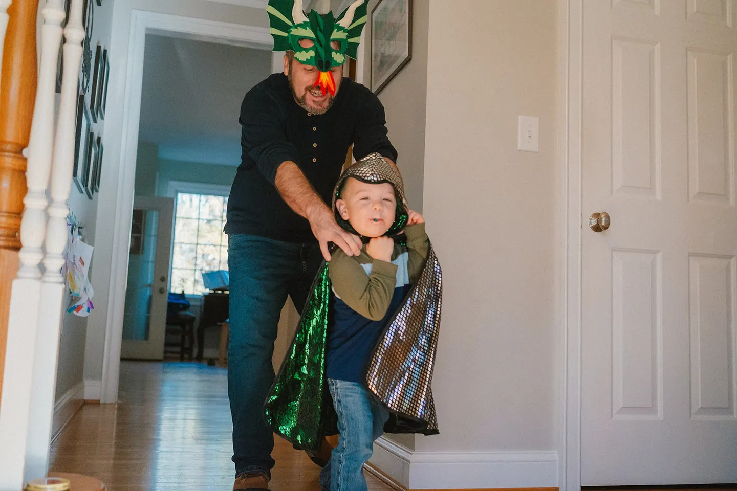 A dad in a dragon mask helps his son into a matching dragon cape in the front hallway.