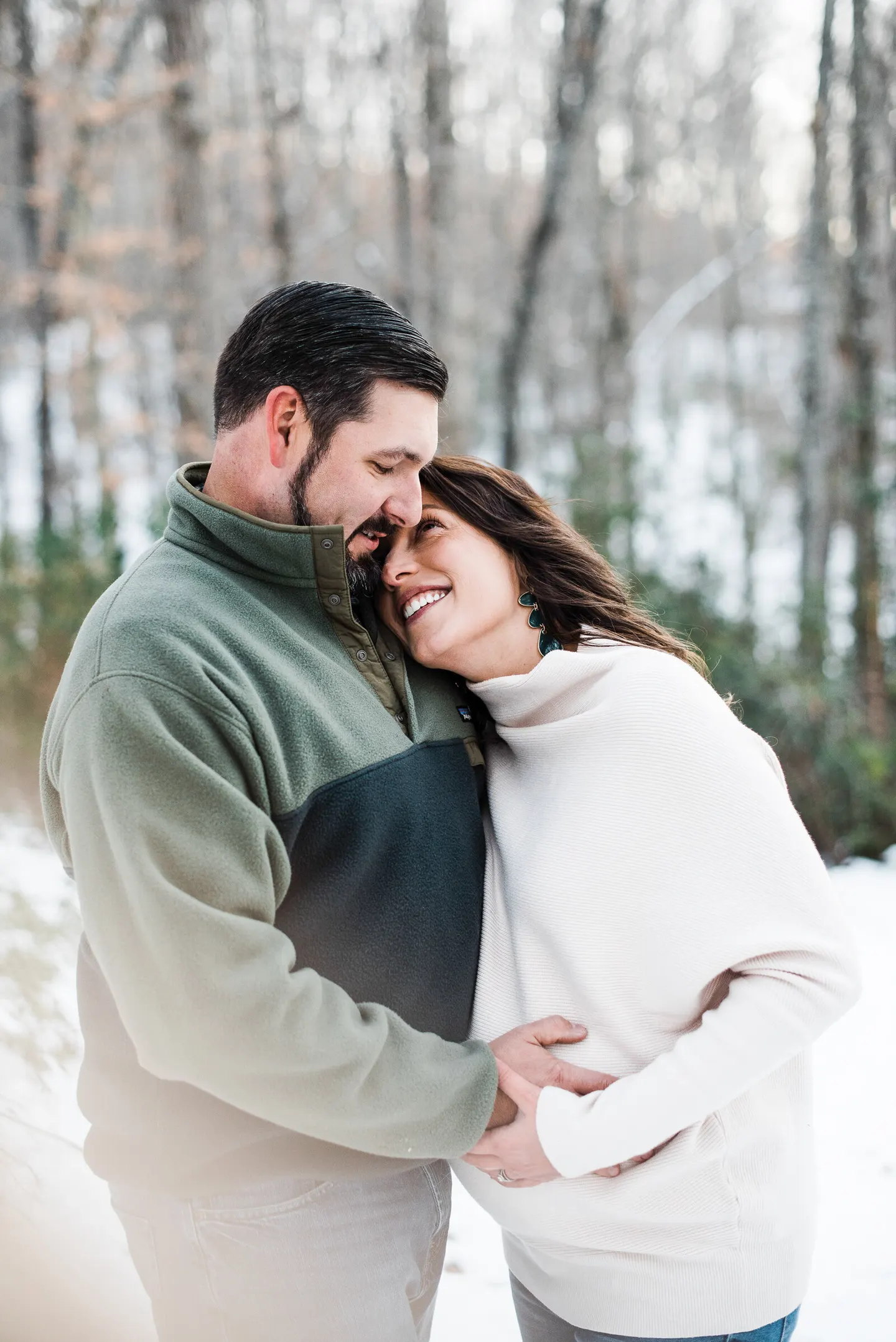 Expectant couple embrace forehead to forehead in a snowy Virginia wood