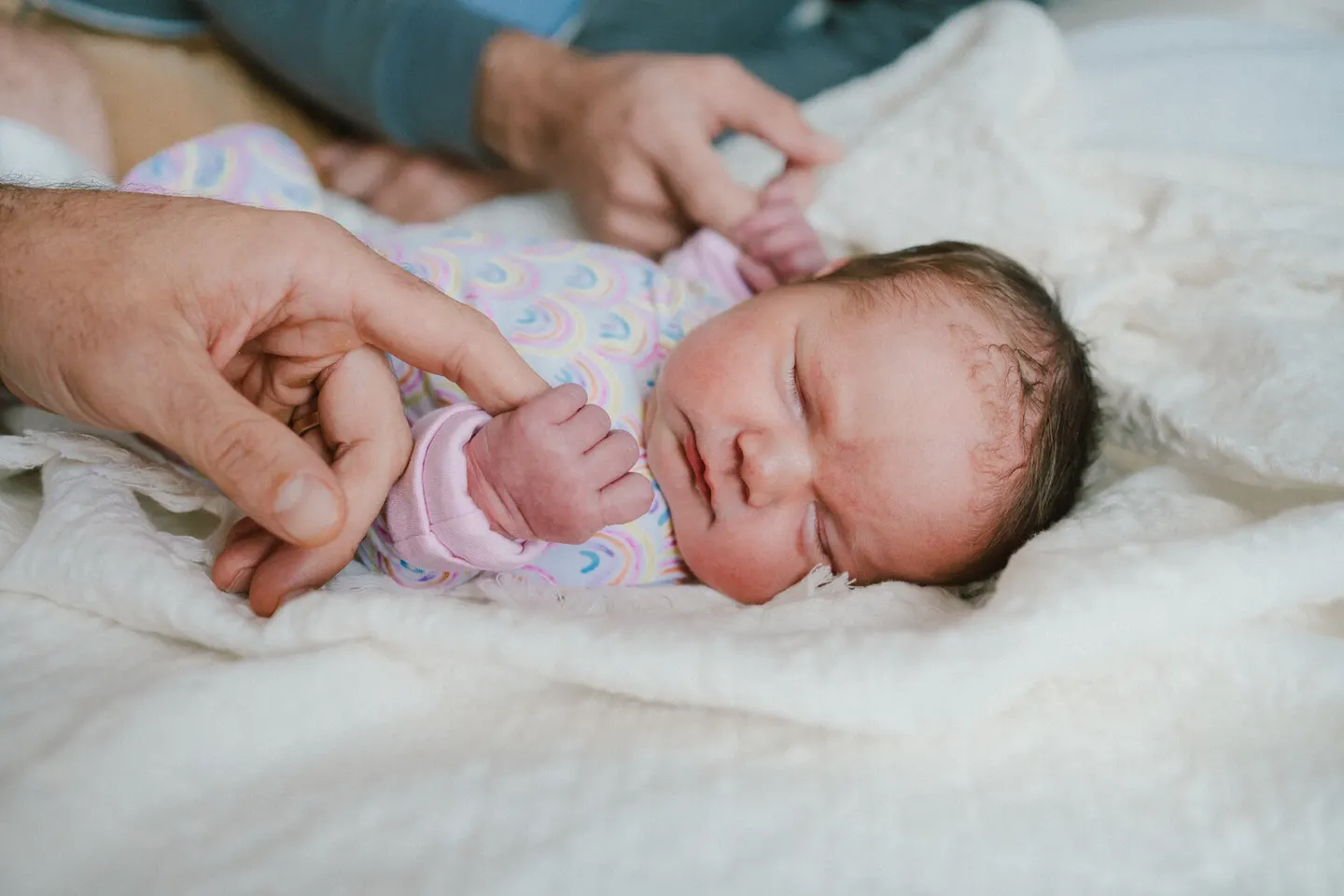 Newborn girl in a rainbow-print onesie sleeps as dad's fingers hold her tiny fist
