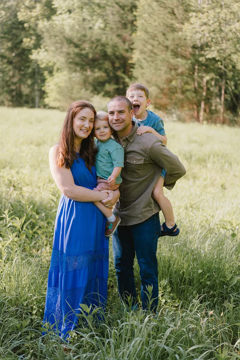Parents hold two young sons close in a lush green field, one boy laughing while piggybacking on dad.