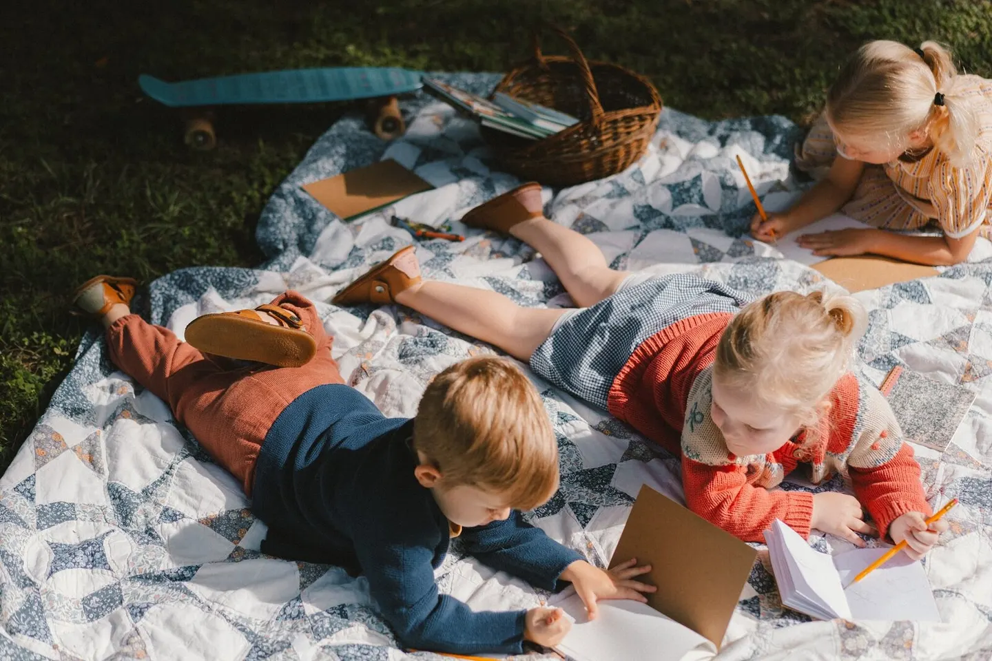 Siblings sprawled on a quilt sketching in notebooks beside a basket and toy skateboard in dappled sunlight
