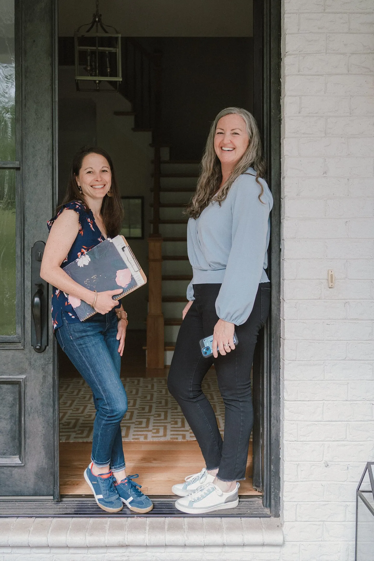 Two real estate agents pose laughing in a home's open front doorway with a clipboard and phone