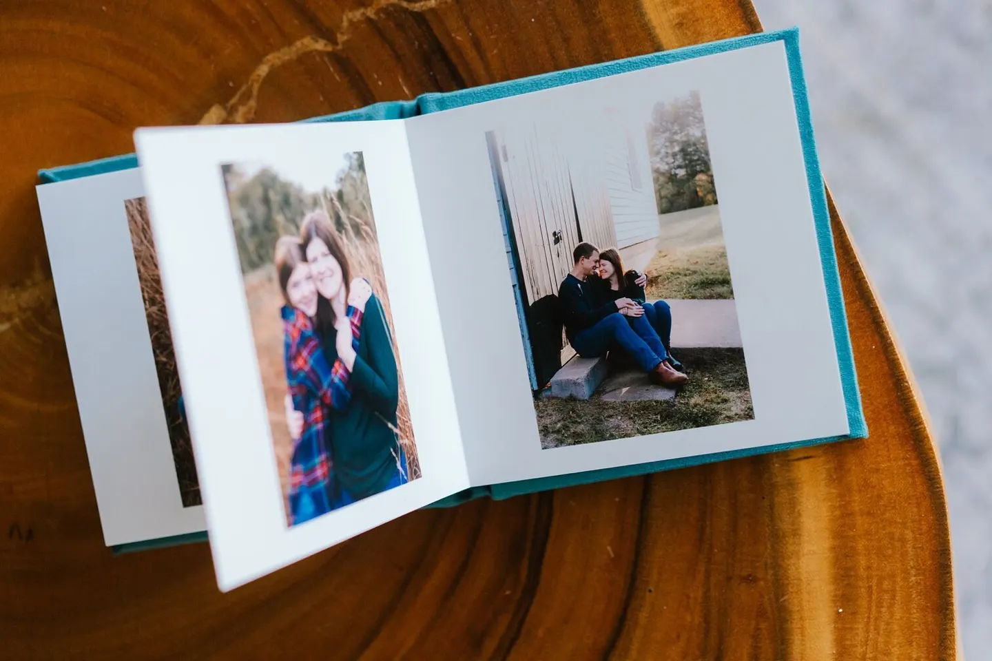 Teal linen album fanned open on a tree-slice table, engagement portraits on facing pages