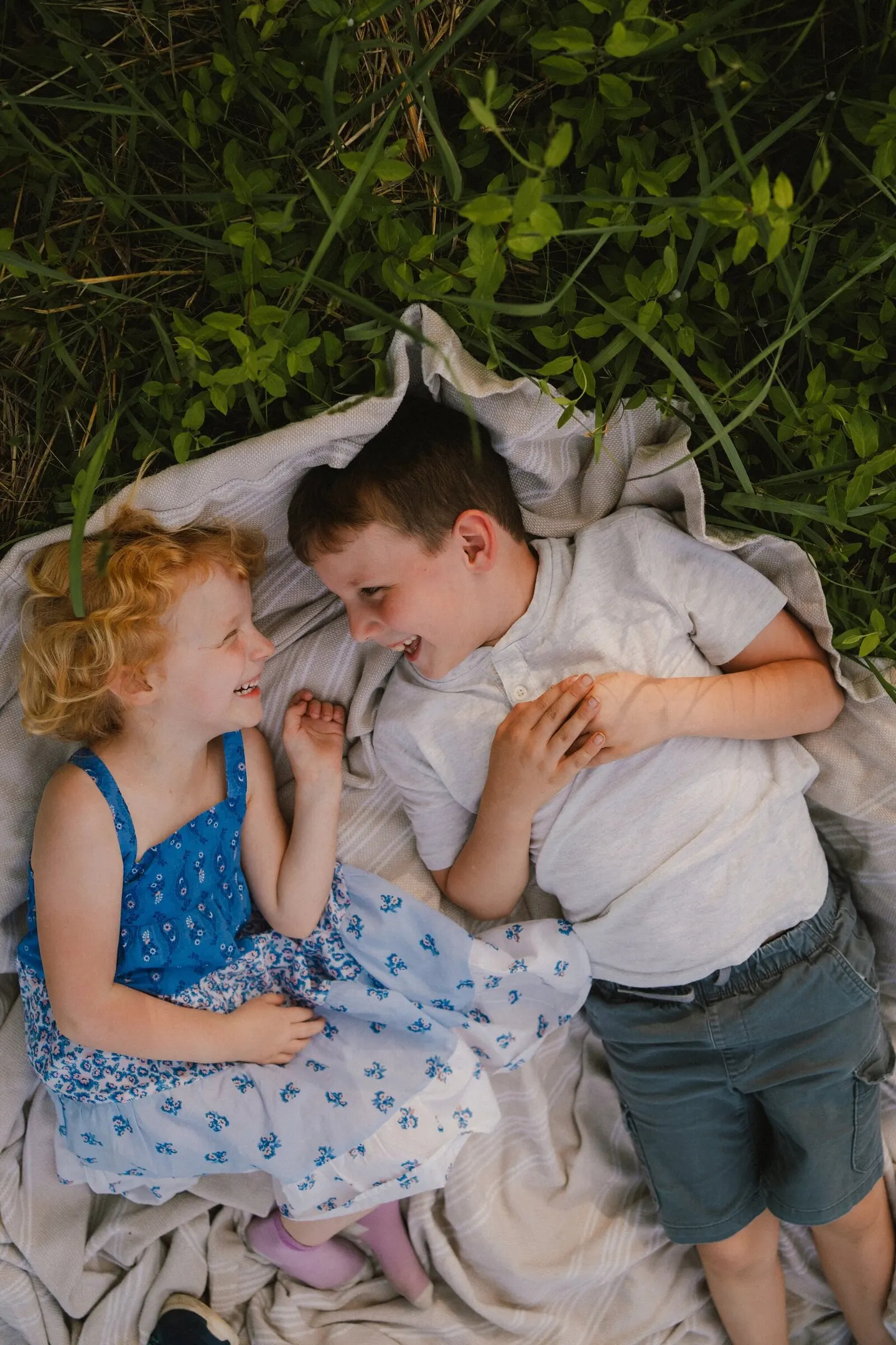 Seen from above, a brother and sister giggle together under a blanket tent in tall backyard grass.
