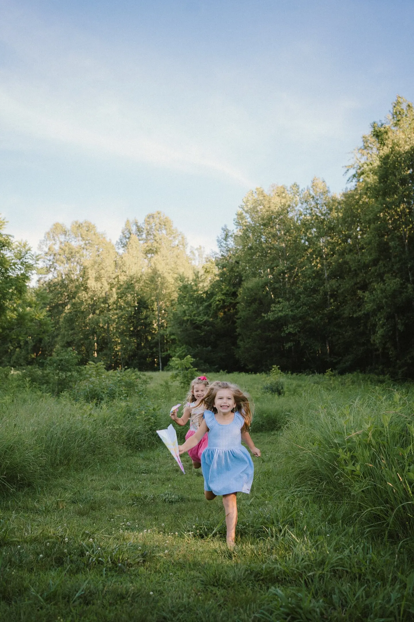 Two barefoot sisters race through tall summer grass holding paper airplanes, hair flying.