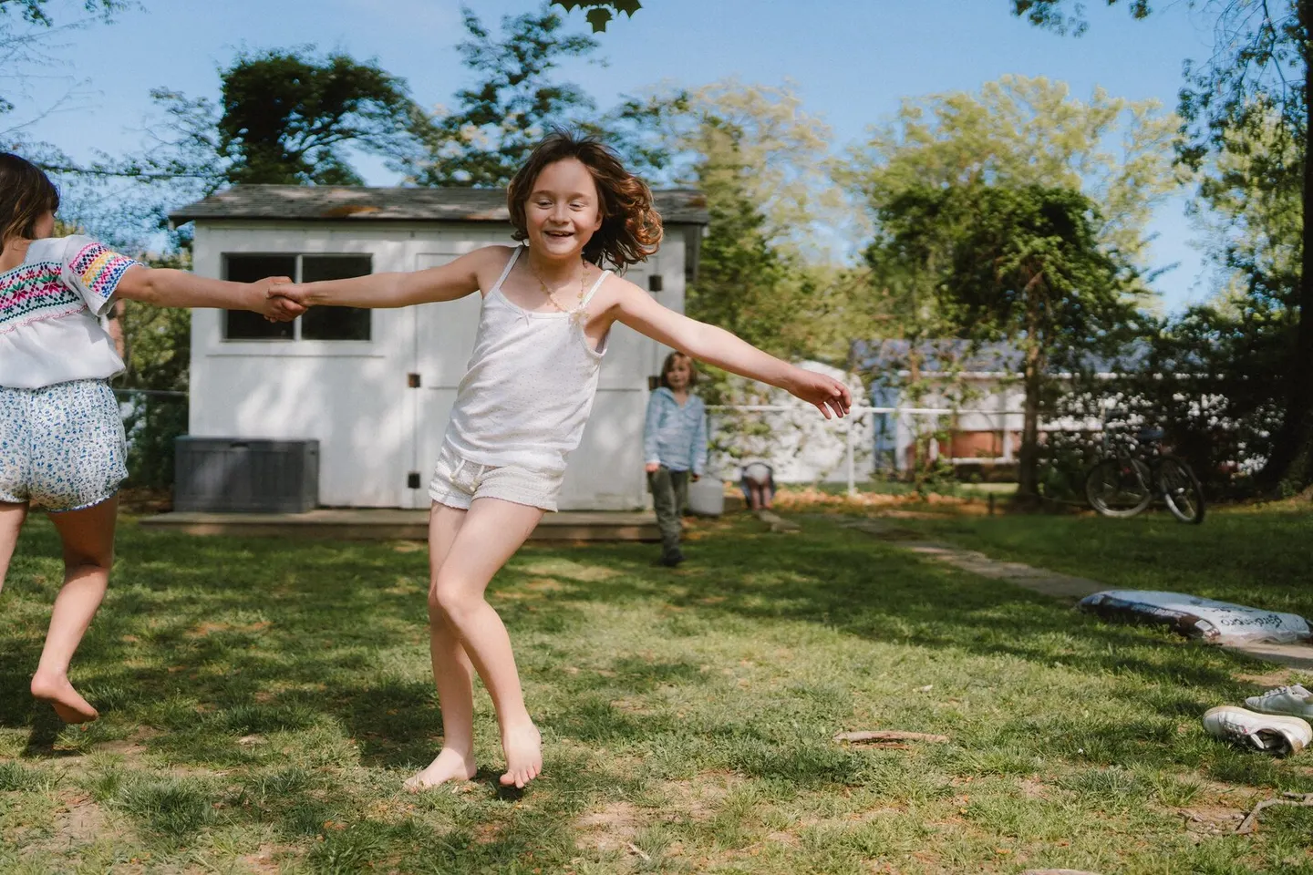A barefoot girl twirls hand-in-hand with a friend across the backyard grass near the garden shed.