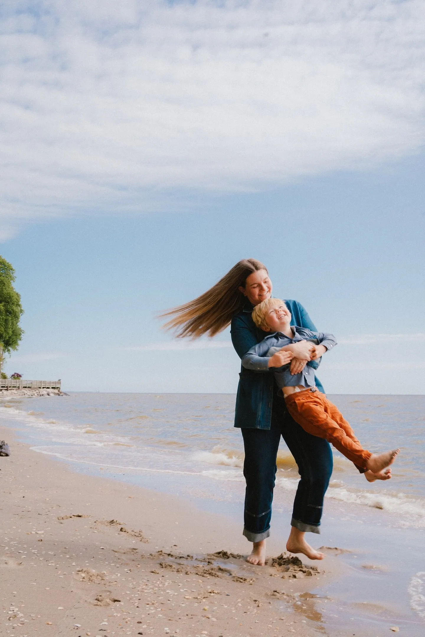 On a windswept beach, a mother swings her son by the arms as waves roll in behind them.