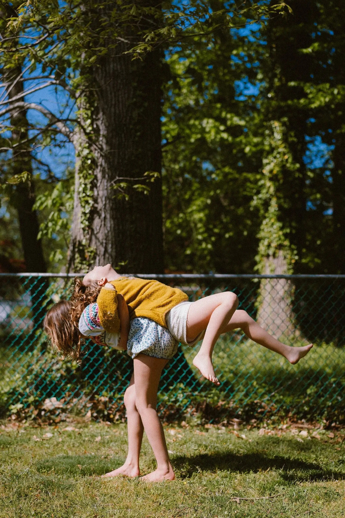 A girl in a yellow sweater arches back playfully while being swung across the lawn.