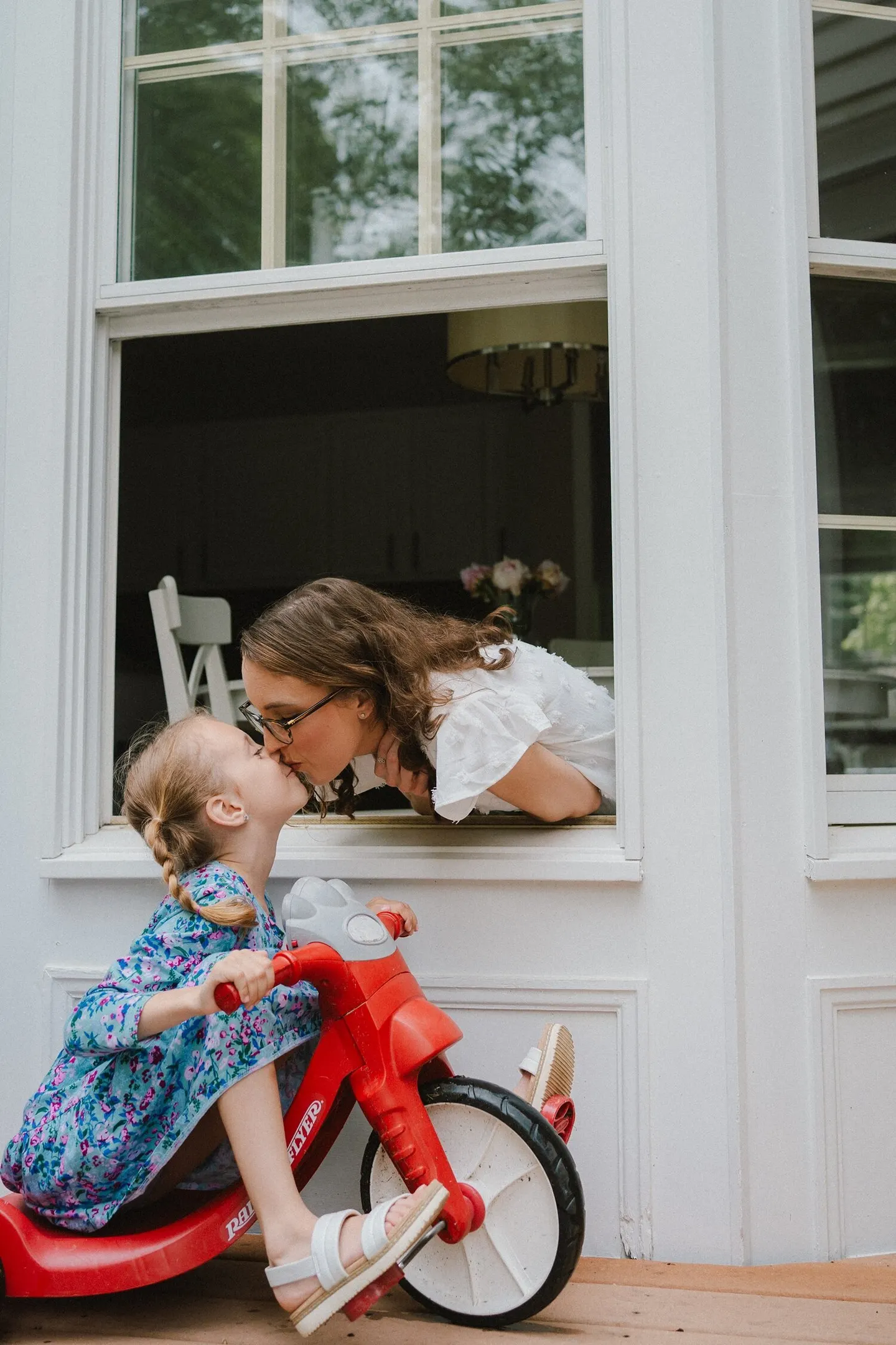 A Charlottesville family photographer captures a mom leaning through an open window to kiss her girl on a tricycle.