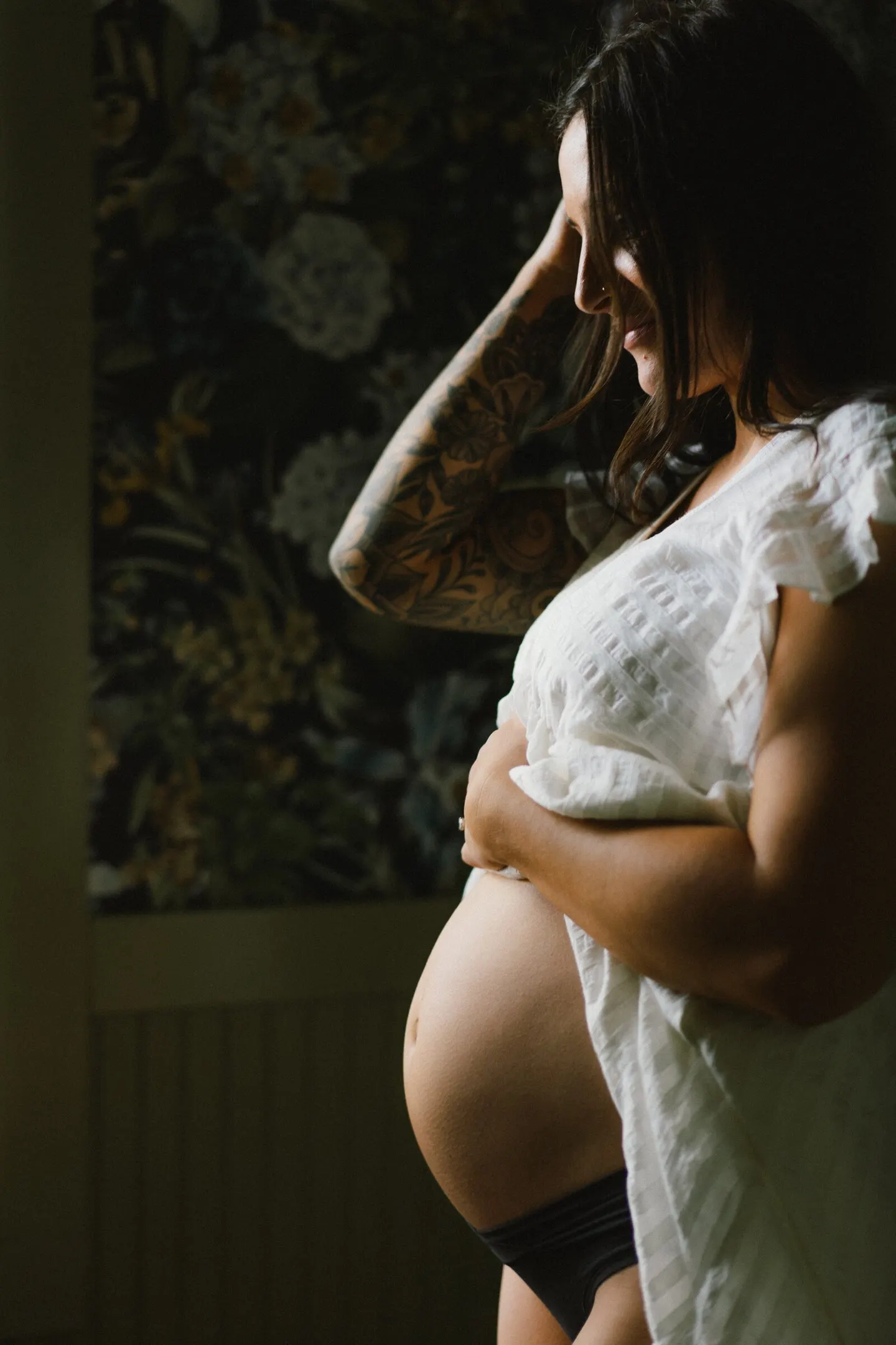 Tattooed pregnant woman gazes down at her bump beside dark floral wallpaper