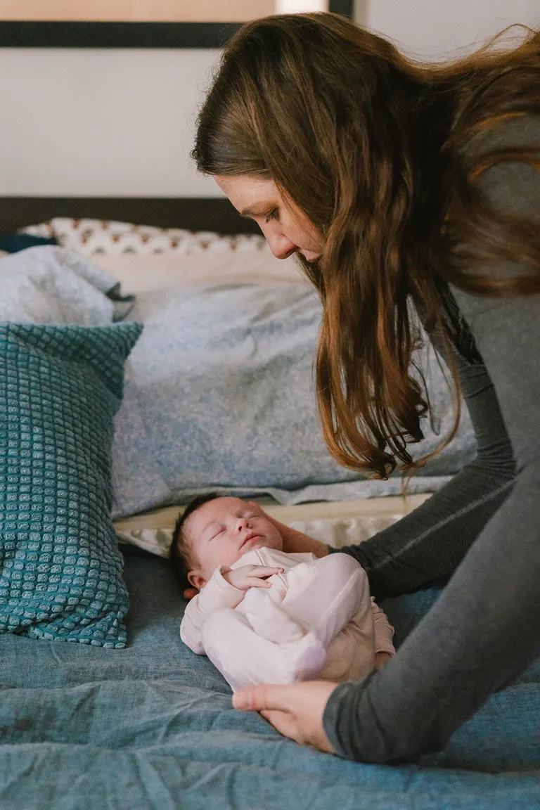 Mother leans over her newborn lying swaddled on a teal-linened bed