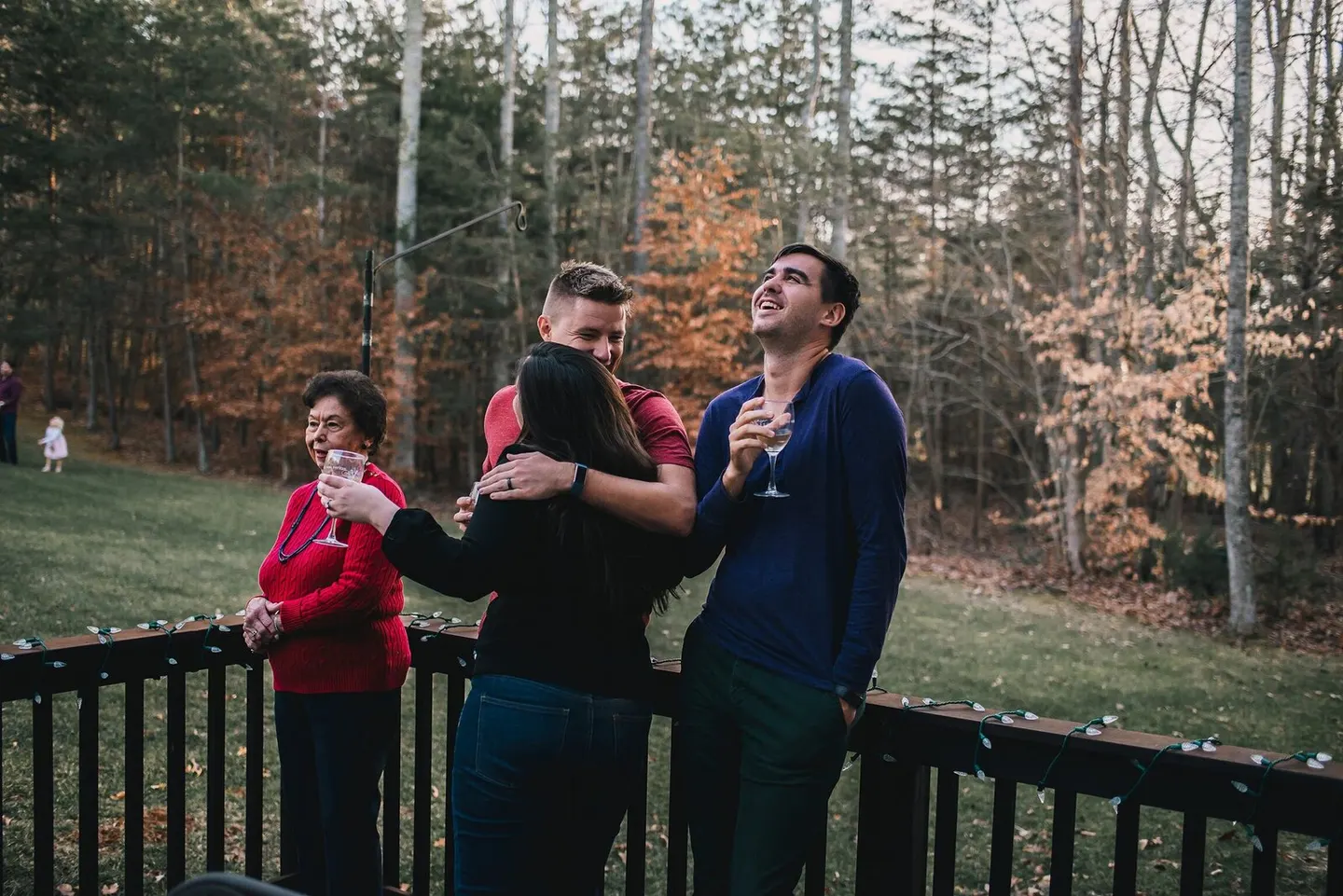 Cousins hug tight on a string-lit deck at dusk while grandma looks on with her wine glass.