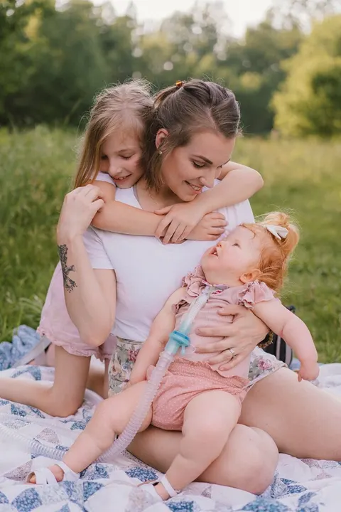 A mother sits in a meadow embracing her two daughters, one snuggled close with her breathing tube in view.