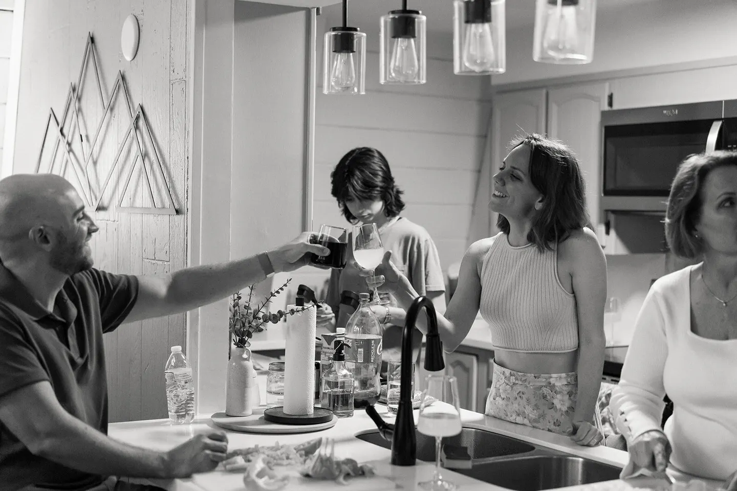 A father and daughter clink glasses in a kitchen while grandma and a teen look on, warm string-light glow.