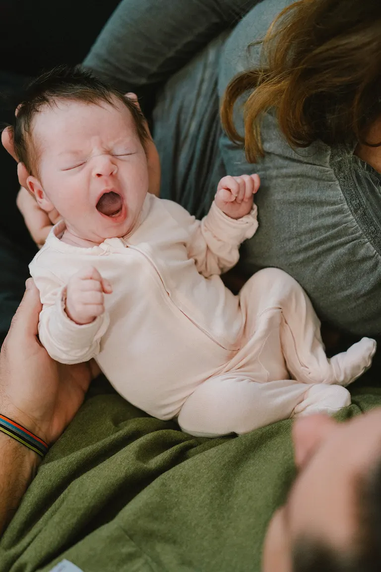 Newborn yawns wide while cradled in a parent's arms on a green blanket