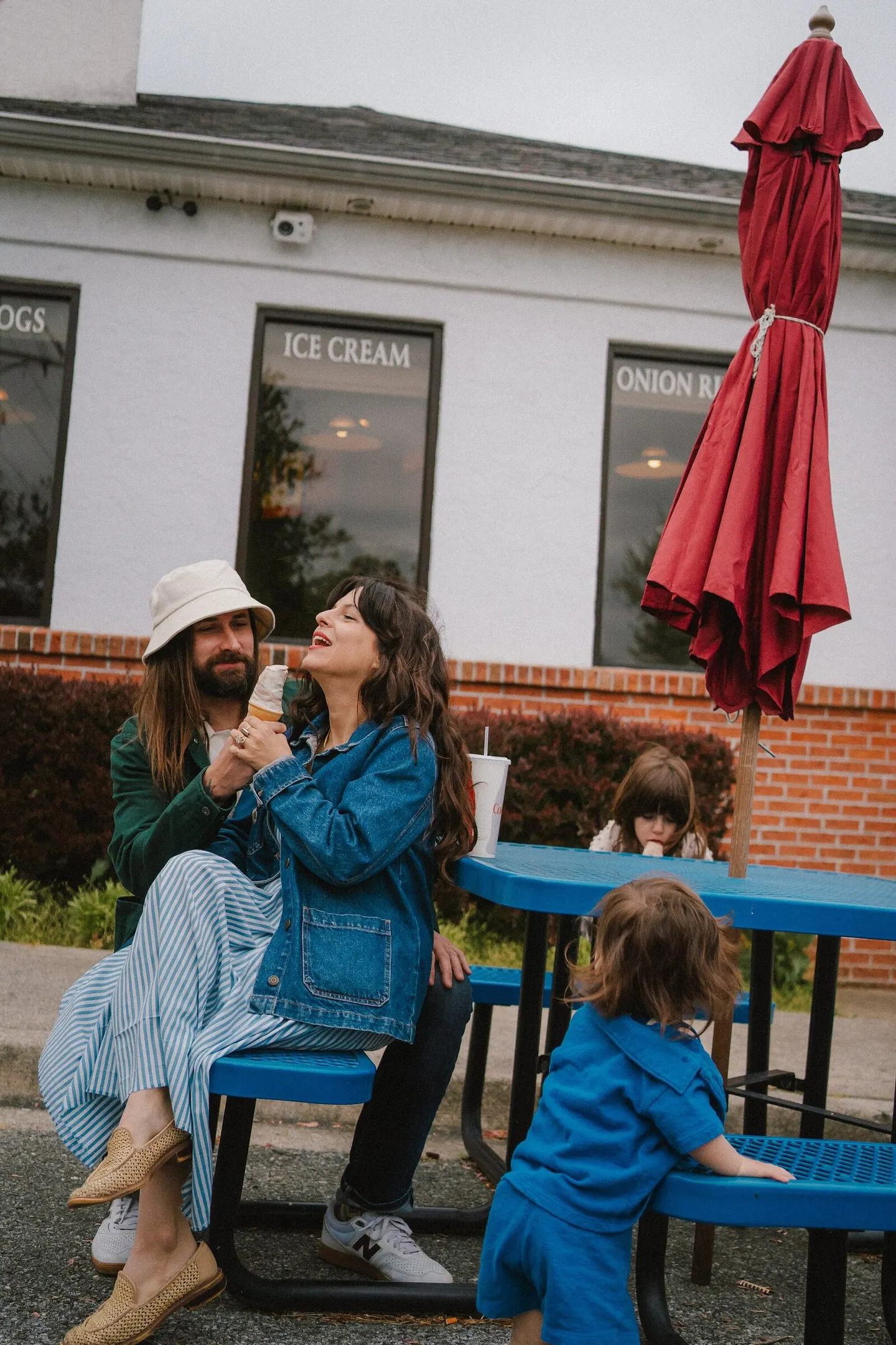 A couple shares an ice cream cone at a picnic table while their daughters look on nearby.