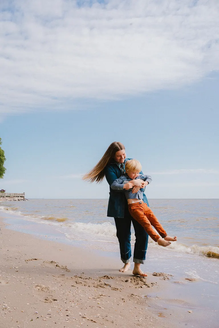 A mother carries her son sideways across the beach as he giggles in her arms.