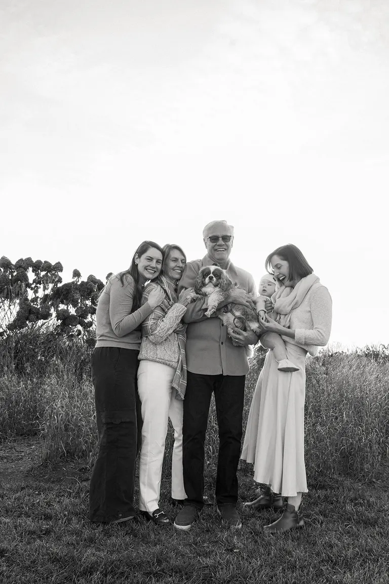 Grandparents, daughters and a baby huddle close in a wildflower field, their spaniel cradled in the middle.