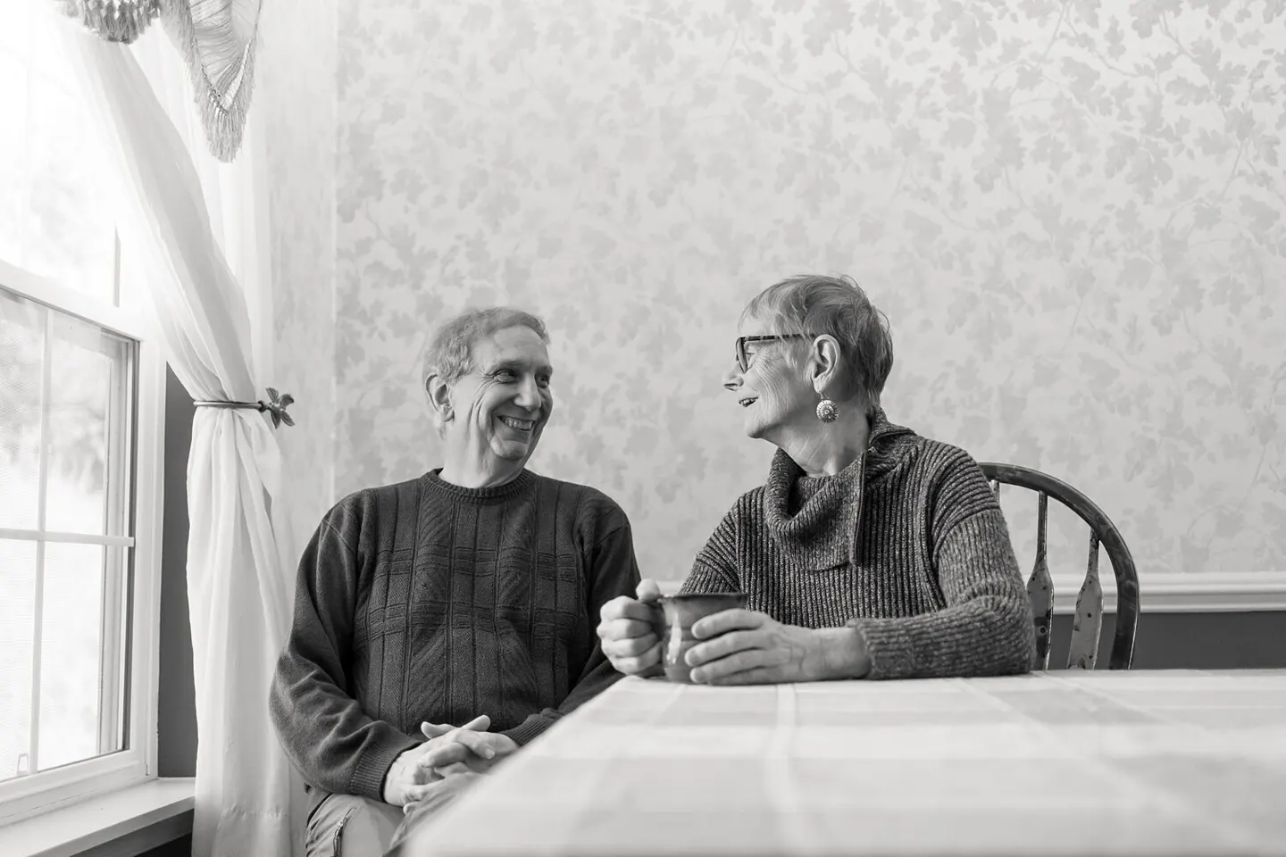 An older couple shares a quiet smile over coffee at a lace-covered kitchen table, black-and-white portrait.