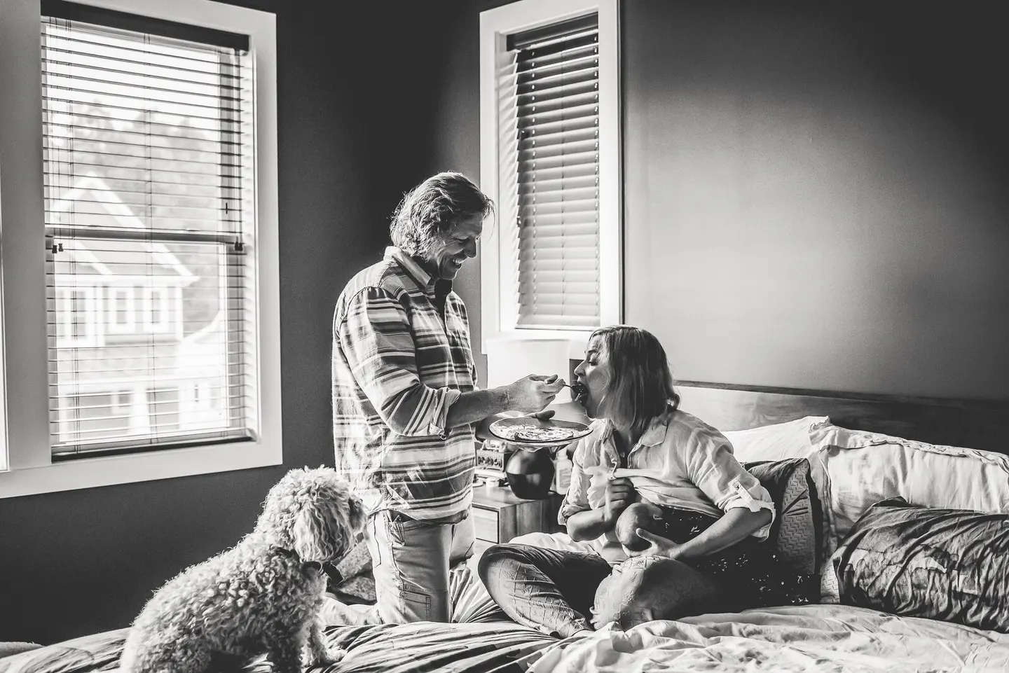 Husband feeds his wife breakfast in bed while she holds their baby and the dog looks on