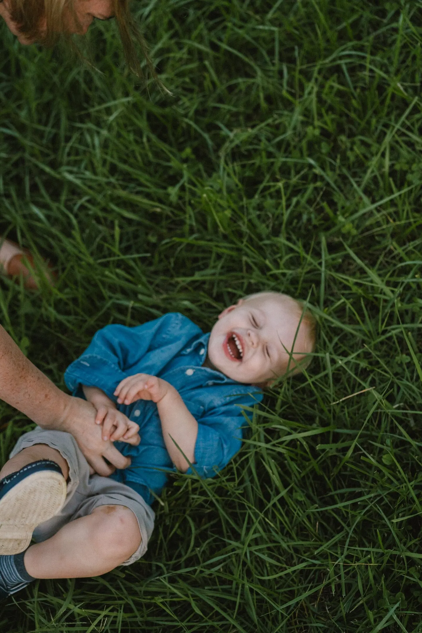 A toddler bursts into laughter lying in deep green grass as his mother's hand steadies him.