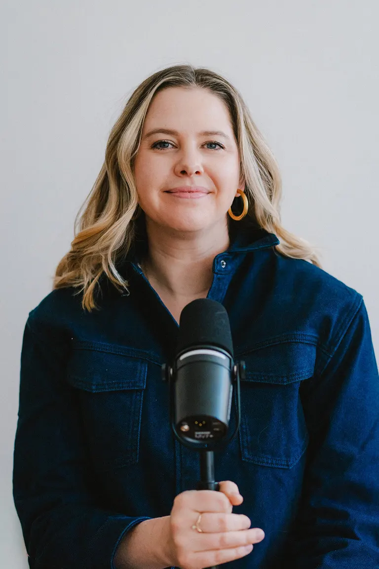 Podcast host in a denim jacket and gold hoops speaks into a microphone against a plain backdrop