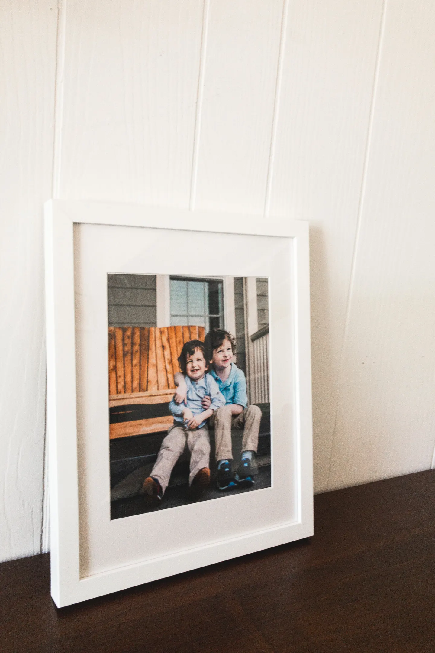 White-matted frame leaning on a shiplap wall, two brothers laughing on a porch bench inside