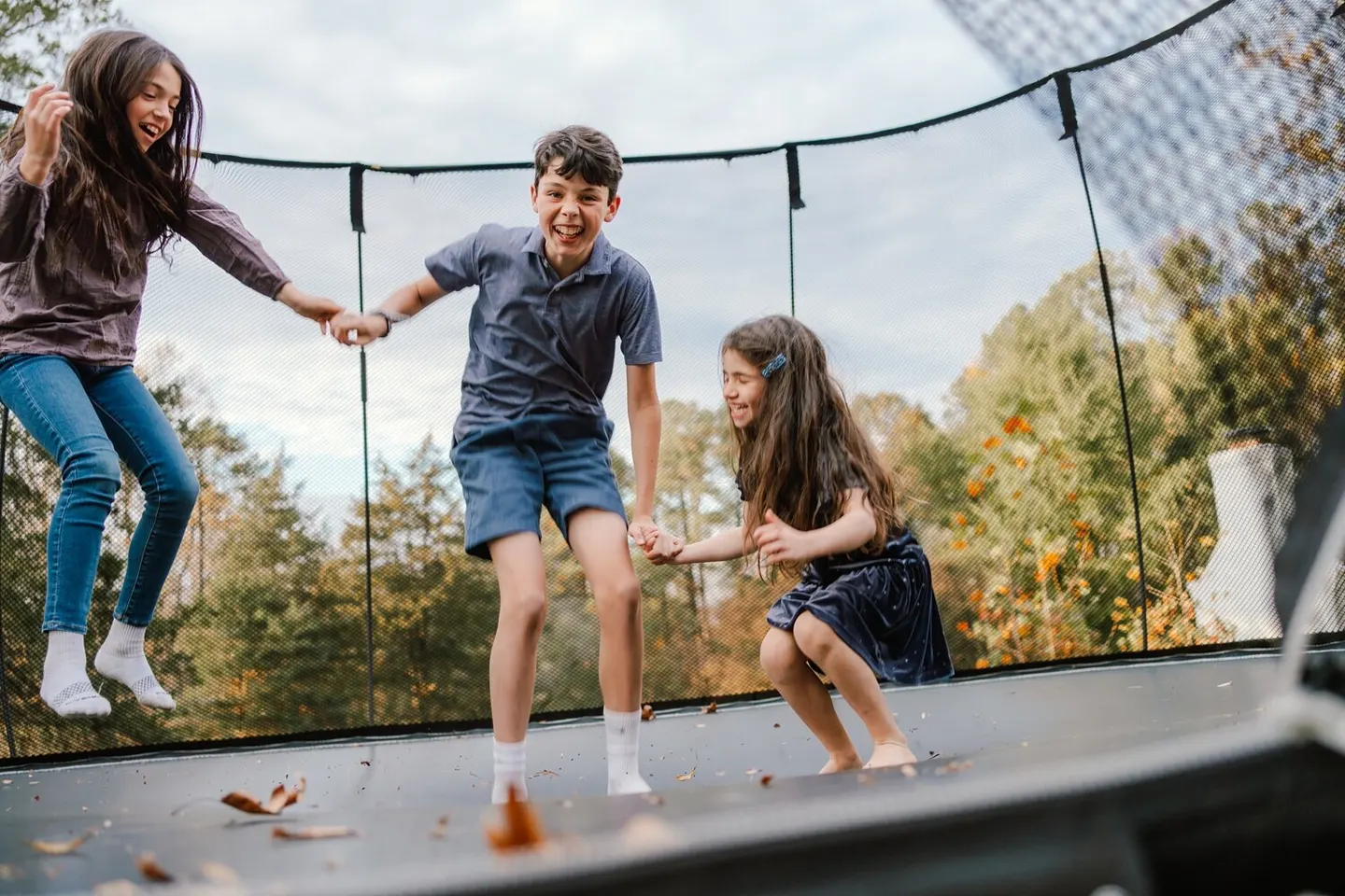 Three kids jump together on a trampoline, hands linked, mid-air and laughing.