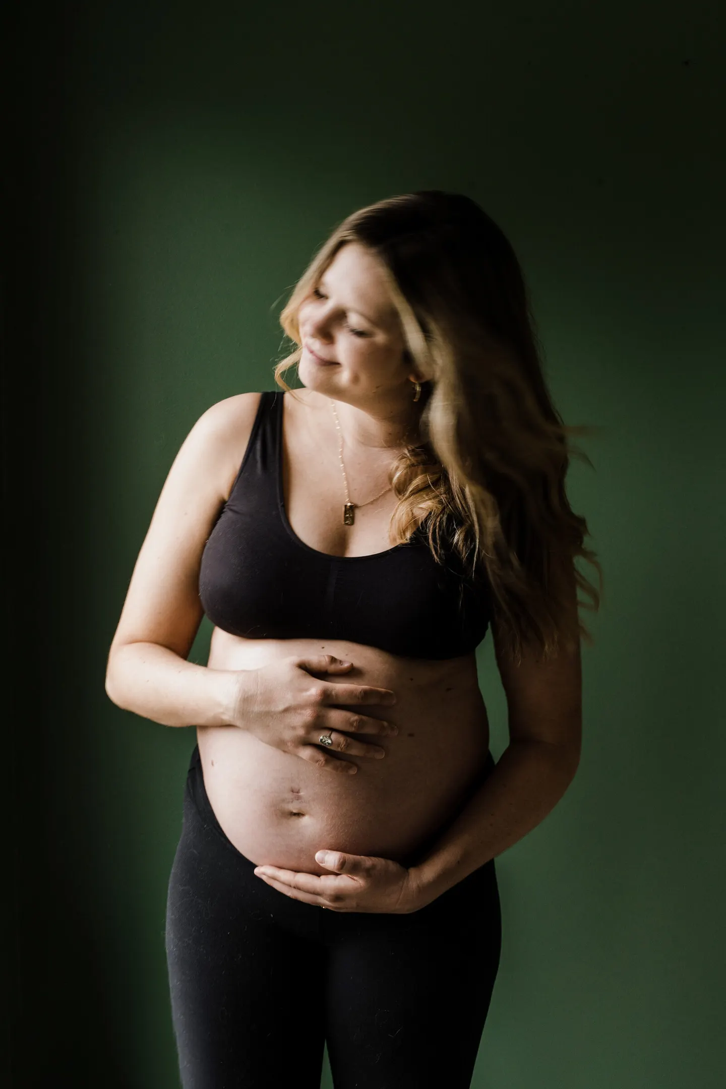 Maternity portrait against a dark green backdrop, hands cradling a bare bump