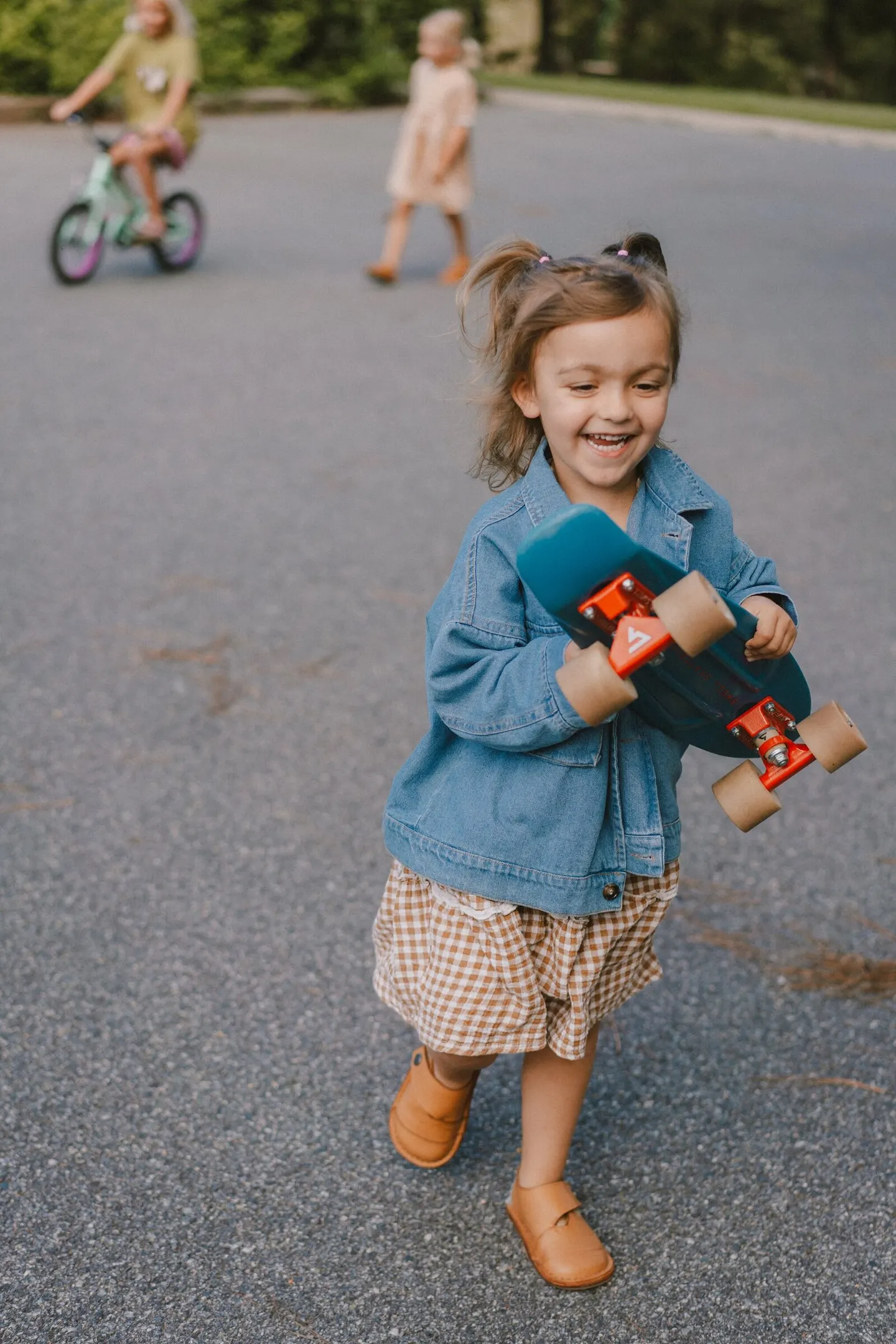 Small girl in gingham skirt and friends bike down a sunny driveway, one clutching a skateboard