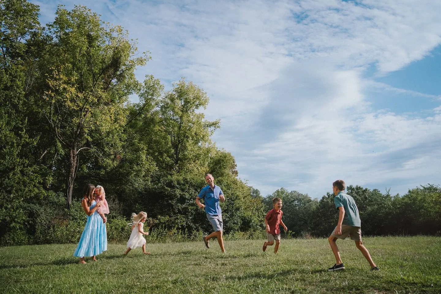 A family scatters across a sunlit meadow, kids sprinting ahead while mom carries a toddler.