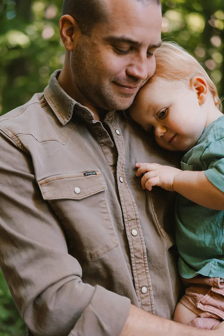 A father rests his cheek against his toddler's head in a quiet, green-lit forest moment.