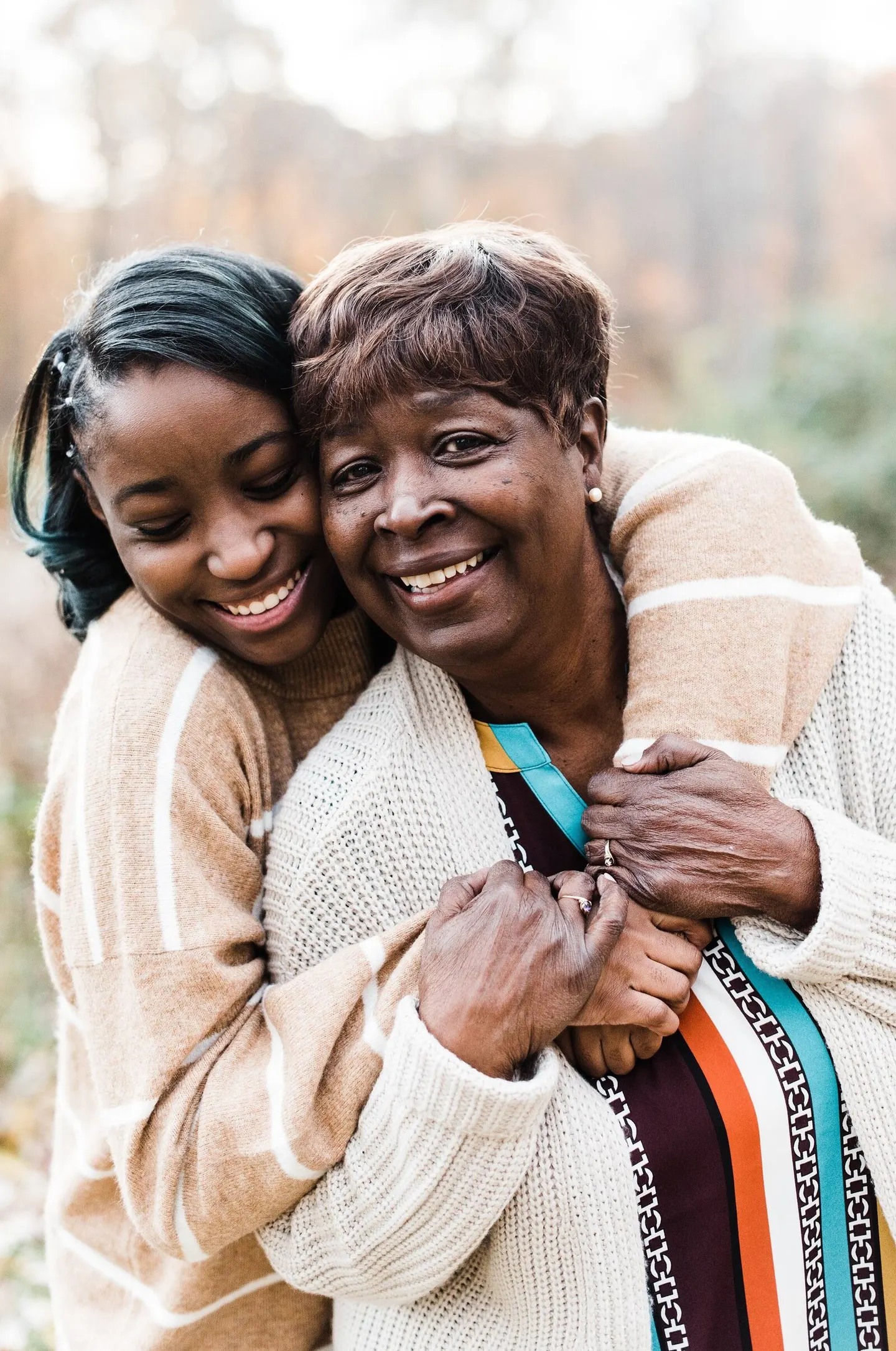 A grandmother and granddaughter share a laughing cheek-to-cheek hug outdoors, hands clasped over her shoulder.