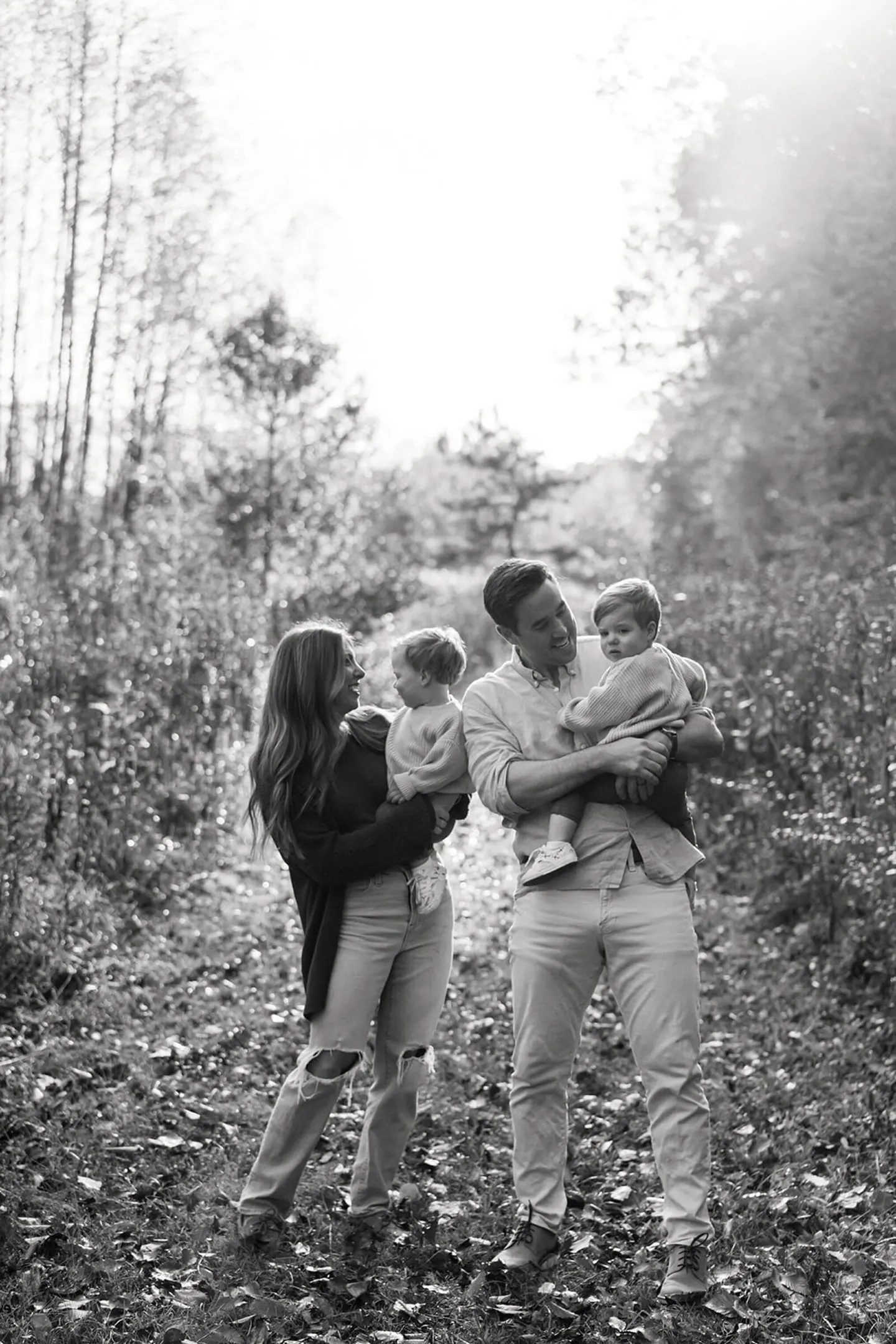 Two parents share a laugh while each holding a toddler on a sunlit, leaf-covered forest trail.