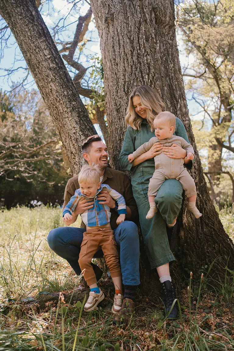 A young family sits nestled among tree roots, each parent balancing a smiling toddler in dappled spring light.