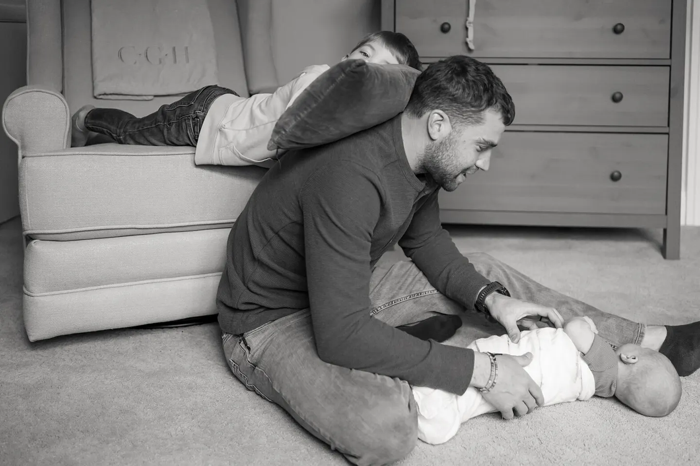 Dad tends to a newborn on the nursery floor while his son drapes over his shoulders