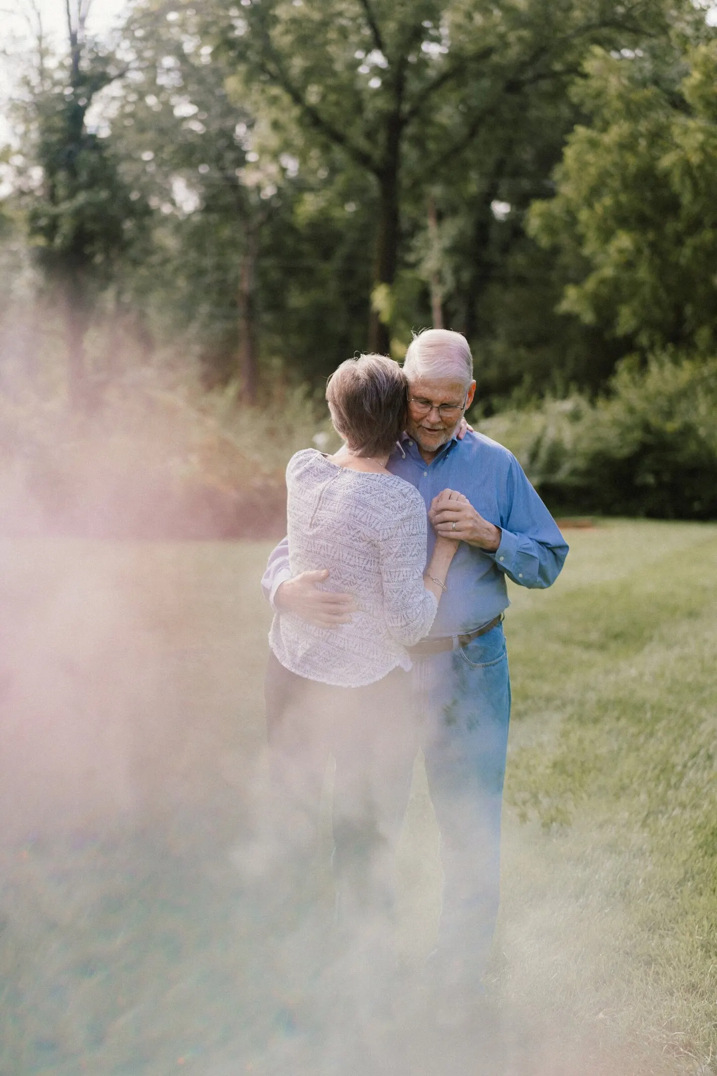 An older couple sways together in a sunlit field, foreheads close, lens flare drifting across the grass.