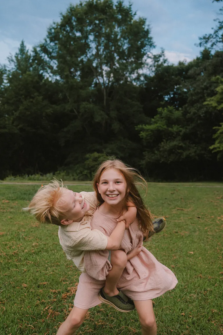 A young girl smiles at the camera while giving her giggling little brother a piggyback ride in an open field.