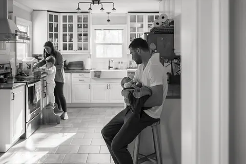 In black and white, a father feeds a newborn while mom and a toddler cook together in the kitchen.