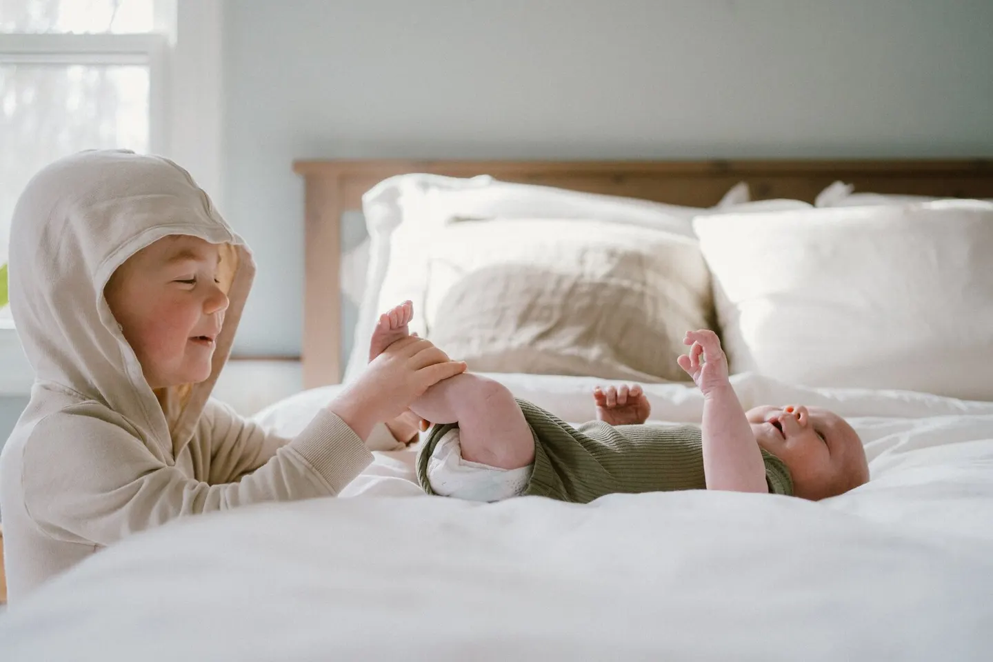 Big brother in a hooded sweatshirt gently holds his newborn sibling's feet on a sunlit bed